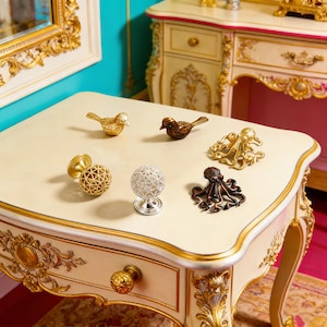 May include: Various decorative metal ornaments, including bird figurines, octopus sculptures, and spherical knobs. The items are in gold, bronze, and silver tones, displayed on a cream-coloured table with ornate gold trim.