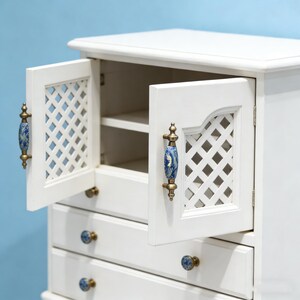 以下が含まれることがあります： 白いキャビネットで、ドアと引き出しが開いています。ドアは格子模様で、青と金の取っ手が付いています。引き出しにはお揃いの青と金のノブが付いています。キャビネットは水色の背景に配置されています。