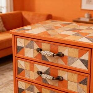 以下が含まれることがあります： オレンジ色の3段引き出しキャビネット。引き出しの前面と上部には幾何学模様が施されています。引き出しの前面には、オレンジ、グレー、クリーム色の三角形のパターンがあります。キャビネットには、花の模様が施された装飾的なハンドルが付いています。