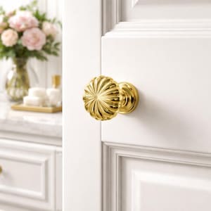 May include: A gold-coloured door knob with a fluted, flower-like design. The knob is mounted on a white door. The door knob is a decorative element, adding a touch of elegance to the door's design.