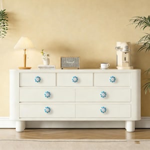 以下が含まれることがあります： 6つの引き出しと曲線的なデザインの白いドレッサー。引き出しの取っ手は青と白の花の形をしています。ドレッサーは、ランプ、スピーカー、コーヒーメーカーのある部屋にあります。