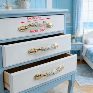 Peut inclure: Commode &agrave; trois tiroirs peinte en blanc et bleu clair. Chaque tiroir est &eacute;quip&eacute; d'une poign&eacute;e dor&eacute;e avec un centre en c&eacute;ramique &agrave; motifs floraux. Les poign&eacute;es mesurent 14,5 cm de long et 3,5 cm de haut.