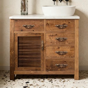 May include: A wooden bathroom vanity unit with a white countertop and a white, oval-shaped sink. The vanity has four drawers and a slatted cabinet door. The hardware is antique brass. A clear glass vase is on the countertop.