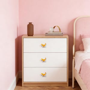 May include: A white three-drawer dresser with orange cat-shaped knobs. The dresser has a light wood top and is set against a pink wall. A brown vase and decorative items sit on top of the dresser.