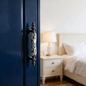 以下が含まれることがあります： 装飾的なハンドルが付いた濃い青色のドア。ハンドルは黒で、白と黒の花柄のデザインです。ドアが開いており、白いナイトスタンド、ランプ、ベッドのある寝室が現れています。