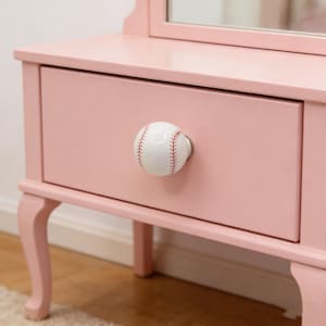 Peut inclure: Une coiffeuse rose avec un tiroir orné d'un bouton en forme de balle de baseball. Le bouton est blanc avec des coutures rouges. La coiffeuse a un miroir et des pieds incurvés, suggérant un style vintage ou décoratif.