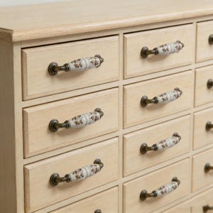 May include: Close-up of a cream-coloured wooden cabinet with multiple small drawers. Each drawer has a decorative handle with a floral pattern in brown and gold. The cabinet's finish is a light, neutral tone.