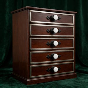 May include: A dark brown wooden chest of drawers with five drawers. Each drawer has a silver-colored metal frame and a white knob. The knobs are centered on decorative black metal accents. The chest is a dark wood color.
