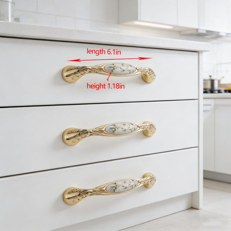 Peut inclure: Poign&eacute;es de meubles dor&eacute;es avec des inserts en c&eacute;ramique florale. Chaque poign&eacute;e mesure 15,5 cm de long et 3 cm de haut. Les poign&eacute;es sont mont&eacute;es sur des tiroirs de meubles blancs, offrant un design classique et &eacute;l&eacute;gant.