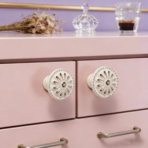 以下が含まれることがあります： ピンク色の引き出し付きのドレッサーのクローズアップ。白い花の形の引き出しノブが2つ付いています。ノブは詳細なデザインで、ピンク色の引き出しに配置されています。ドレッサーにはシルバー色のハンドルも付いています。