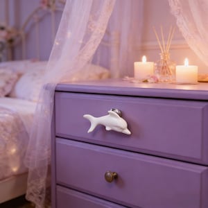May include: A lavender nightstand with a white dolphin-shaped drawer pull. The nightstand has two drawers with a brass knob on the bottom drawer. Two lit candles and a glass diffuser sit on top of the nightstand.