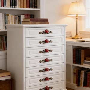 以下が含まれることがあります： 真鍮色の装飾的なハンドルと赤いノブが付いた、白い6段のチェスト。本棚とランプのある部屋にあり、図書館や書斎を連想させます。2冊の本がチェストの上に置かれています。