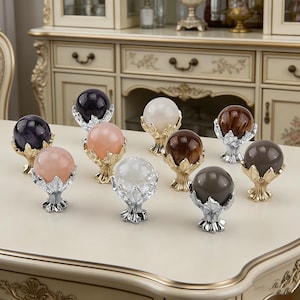 Op de afbeelding: Een verzameling decoratieve bollen in verschillende kleuren, waaronder zwart, roze, wit en bruin. Elke bol wordt vastgehouden door een metalen, handvormige standaard. De bollen zijn gerangschikt op een lichtgekleurde tafel, met een kast op de achtergrond.