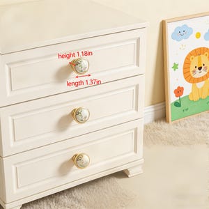 Peut inclure: Un meuble &agrave; trois tiroirs cr&egrave;me avec des boutons &agrave; motifs floraux. Les boutons sont dor&eacute;s avec un motif floral blanc et vert. La hauteur du bouton est de 3 cm et la longueur de 3,5 cm. Une image encadr&eacute;e d'un lion de dessin anim&eacute; est &agrave; droite.
