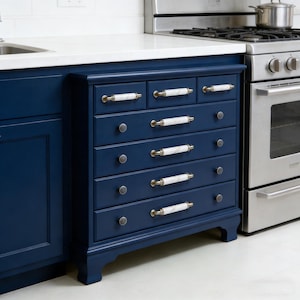 以下が含まれることがあります： ネイビーブルーのキャビネット。7つの引き出しと、お揃いのドアがあります。引き出しには、白い大理石風のハンドルとゴールドカラーのアクセントが付いています。キャビネットはステンレス製のストーブと白いカウンタートップの隣にあります。
