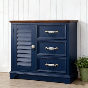 以下が含まれることがあります： 濃い茶色の天板が付いたネイビーブルーのキャビネット。キャビネットの左側にはルーバー式のドアがあり、右側には3つの引き出しがあります。引き出しには、金色のアクセントが付いた花柄のハンドルが付いています。右側には小さな鉢植えがあります。