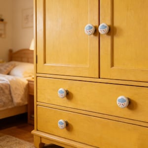 May include: A yellow wooden cabinet with white knobs featuring a blue cloud and rain design. The cabinet has two doors and two drawers. The knobs are round and add a decorative touch to the furniture.