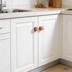 以下が含まれることがあります： ピンクとゴールドのキャビネットノブが付いた白いキッチンキャビネットのクローズアップ。ノブは幾何学的なデザインで、白いキャビネットのドアに取り付けられています。キッチンはモダンなデザインです。