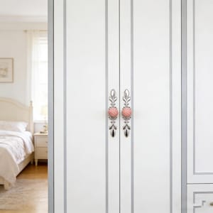 以下が含まれることがあります： 白いクローゼットのドア、シルバーのトリムと装飾的なピンクとシルバーのノブ。ノブは花の模様とテクスチャ加工されたピンクの中央部があります。クローゼットのドアは、ベッドとナイトスタンドが見える寝室にあります。