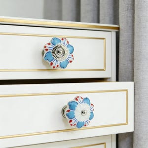 May include: Close-up of a white drawer with a gold trim and a floral ceramic knob. The knob features a white base with blue and red floral details and a silver metal center. The drawer is part of a white cabinet.