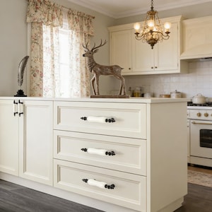 May include: A cream-colored kitchen island with three drawers, each featuring white and black decorative handles. A decorative deer statue stands on top of the island. A chandelier hangs above the island, and a floral curtain is visible in the background.