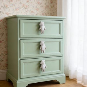 Peut inclure: Commode à trois tiroirs peinte en vert clair. Chaque tiroir est orné d'un bouton en forme d'hippopotame gris. Le meuble est placé devant un papier peint floral et un rideau blanc.