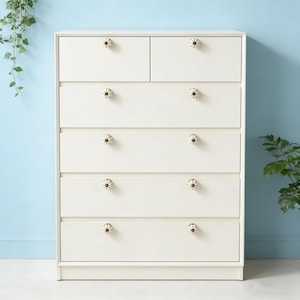 以下が含まれることがあります： 6つの引き出しと2つの小さな上部引き出しが付いた、オフホワイトのドレッサー。各引き出しには、花型の装飾的なノブが付いています。ドレッサーは、背景に緑のある水色の壁に設置されています。