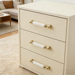 Puede incluir: Cómoda crema de tres cajones con herrajes dorados y blancos. Cada cajón tiene un tirador de cerámica blanca con detalles dorados. La cómoda está sobre un fondo neutro, lo que sugiere un dormitorio o sala de estar.
