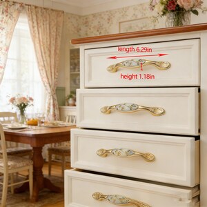 Peut inclure: Une commode cr&egrave;me avec quatre tiroirs, chacun orn&eacute; d'une poign&eacute;e dor&eacute;e avec un motif floral. Les poign&eacute;es mesurent 16 cm de long et 3 cm de haut. La commode est plac&eacute;e devant une salle &agrave; manger.