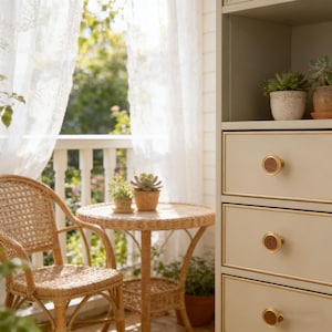 May include: A sunlit outdoor scene features a wicker chair and table, with potted plants. A cabinet with drawers and gold trim is on the right. Sheer white curtains frame a view of greenery, creating a cozy, inviting space.