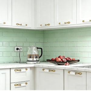 May include: A kitchen with white cabinets and gold and white handles. A glass electric kettle is on the counter, and a wooden bowl holds pomegranates. The backsplash is light green brick.