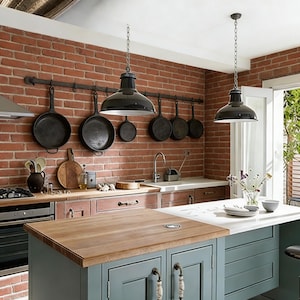 May include: A kitchen with a brick wall, featuring hanging black pendant lights and cookware. A wooden-topped island with blue cabinets and decorative handles is in the foreground. The kitchen has a rustic, farmhouse style.