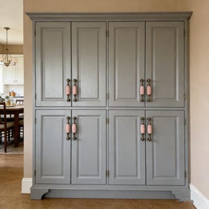 May include: A large, grey cabinet with eight doors and decorative pink and brass handles. The cabinet is a built-in unit with a classic design, suitable for storage in a kitchen or dining area. The cabinet is painted in a neutral grey colour.