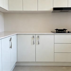 May include: A modern kitchen with white cabinets and countertops. The cabinet doors feature unique fork-shaped handles. A black stovetop and range hood are visible in the background, creating a clean and minimalist aesthetic.