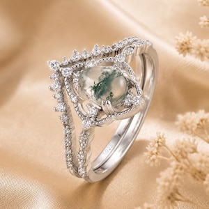 Op de afbeelding: Een zilveren ringenset met een centrale ovale mosagaatsteen met groene insluitingen, omringd door een decoratief frame van kleine heldere steentjes. De ring is gemarkeerd met S925 en bevat een V-vormige band versierd met heldere steentjes.