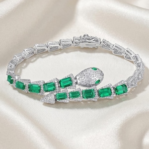 May include: A silver snake bracelet with emerald green rectangular stones and small clear stones. The snake head is detailed with small clear stones and two emerald green stones for eyes. The bracelet is on a white satin surface.
