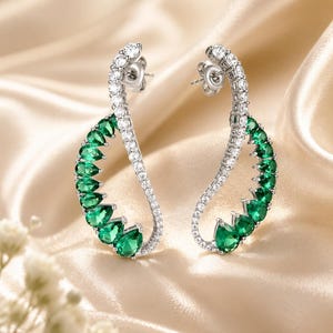 May include: A pair of silver-toned earrings featuring a curved design. Each earring is adorned with a row of small, clear, round-cut stones along the outer edge and a series of larger, pear-shaped, vibrant green stones along the inner curve.