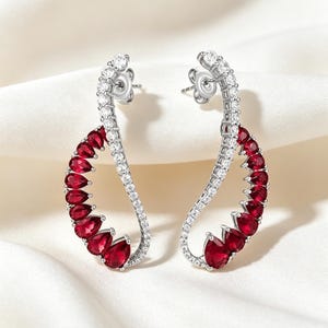 May include: A pair of silver-toned earrings featuring a curved, S-shaped design. Each earring is adorned with a row of small, clear, round-cut stones along the outer edge and a series of deep red, pear-shaped stones along the inner curve.