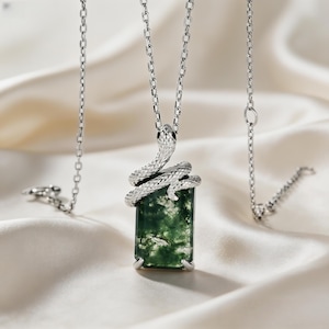 Op de afbeelding: Zilverkleurige ketting met een rechthoekige hanger van mosgroene steen. Een gedetailleerde slang wikkelt zich om de steen. De ketting hangt aan een fijne ketting, op een witte, zijdeachtige achtergrond.