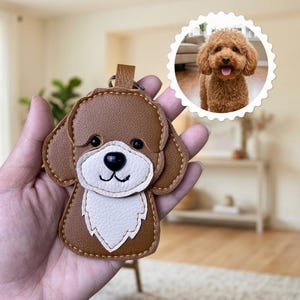 Peut inclure: Un porte-clés en similicuir marron et blanc en forme de chien avec un motif cousu, un nez et des yeux noirs. Une image circulaire insérée montre une photographie d'un chien brun aux poils bouclés avec la langue sortie, assorti au porte-clés.
