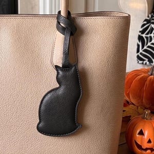 May include: A beige textured tote bag featuring a black leather cat-shaped bag charm attached to the handle. The charm is a silhouette of a sitting cat with a pebbled texture and stitched edges, hanging from a thin black leather strap.