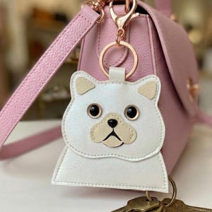 Könnte beinhalten: Ein weißer, katzenförmiger Schlüsselanhänger mit schwarzen Augen und beigen Akzenten hängt an einer rosa Handtasche. Der Schlüsselanhänger hat einen goldfarbenen Ring. Die Handtasche hat einen passenden rosa Riemen.