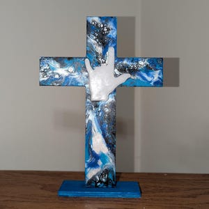 Puede incluir: Una cruz de madera con un diseño abstracto azul, blanco y negro. Una mano blanca con el signo de "Te amo" está fijada en la parte delantera. La cruz está montada sobre una base azul, adecuada para exhibir en una estantería o mesa.