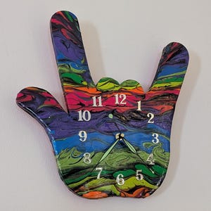 Puede incluir: Un reloj de pared colorido con forma de mano haciendo el signo de 'Te amo'. La esfera del reloj presenta un diseño de pintura vibrante y arremolinado en tonos de morado, verde, naranja y azul. Los números de las horas son plateados y las manecillas son verdes.