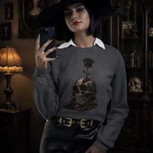 Könnte beinhalten: Dunkelgraues Sweatshirt mit einem Totenkopf- und Rosen-Motiv. Das Model trägt einen schwarzen Hexenhut mit goldener Schnalle und schwarze Lederhose. Der Hintergrund zeigt antike Möbel und gerahmte Porträts.
