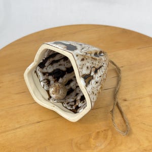 Op de afbeelding: Een keramische bel met een gespikkeld bruin en wit patroon, een beige interieur en een kleine, kogelvormige klepel. De bel heeft een touw van natuurlijke vezels om op te hangen. De bel ligt op een houten oppervlak.