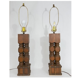 Può includere: Coppia di lampade da tavolo vintage in legno. Ogni lampada presenta una base in legno marrone scuro impilata con un design decorativo. Le lampade hanno un collo color ottone e una lampadina. Le lampade sono alte circa 76 cm.