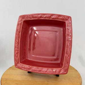 Könnte beinhalten: Ein quadratischer, roter Keramik-Servierteller mit einem Blattmuster am Rand. Die Schale hat eine glänzende Oberfläche und wird auf einer Holzoberfläche präsentiert. Die Schale hat einen leuchtenden Rotton.