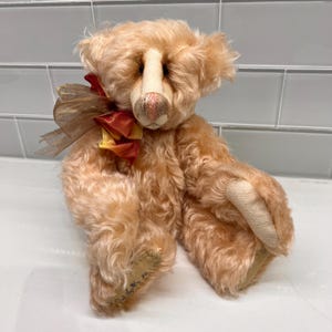 May include: A vintage-style teddy bear with peach-colored fur and a cream-colored snout and paws. The bear has a red and gold ribbon around its neck. The bear is sitting down and has a tag on its foot.