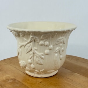 Puede incluir: Una maceta de cerámica de color crema con un diseño en relieve de ramas de cerezo con hojas y frutos. El recipiente tiene un borde ancho y acampanado, con una superficie texturizada y un acabado brillante que resalta los detalles.